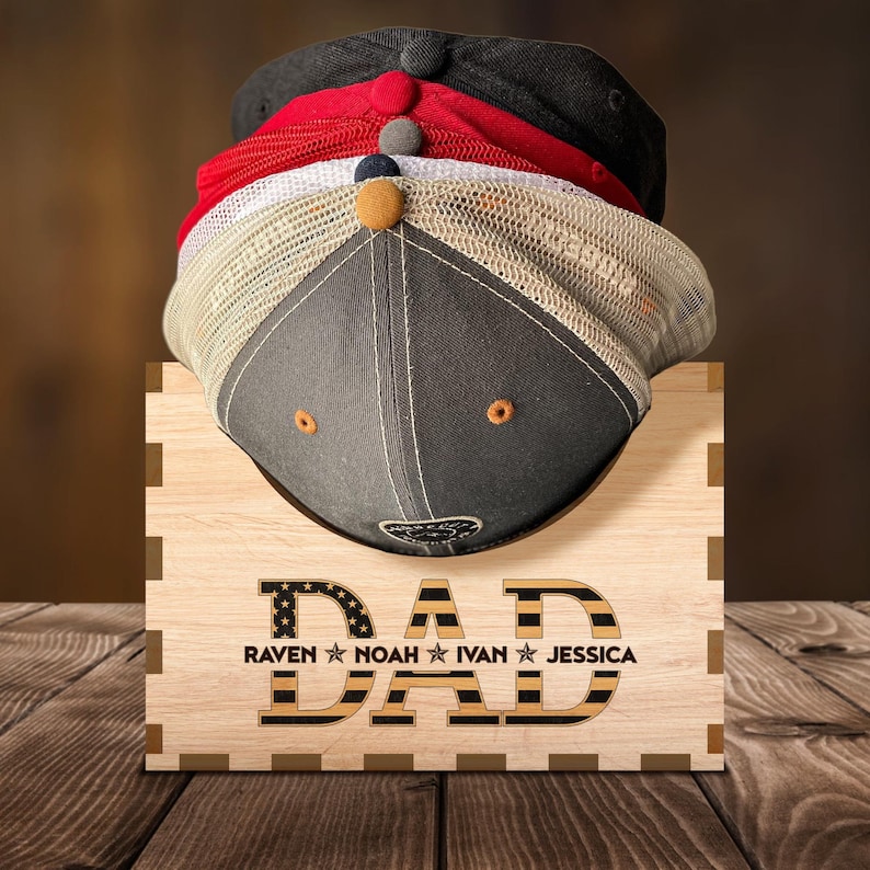Puede incluir: Estante de sombreros de madera con la palabra "DAD" en letras negras con un patr&oacute;n de bandera estadounidense rojo, blanco y azul. Los nombres "RAVEN", "NOAH", "IVAN" y "JESSICA" est&aacute;n escritos debajo de la palabra "DAD".