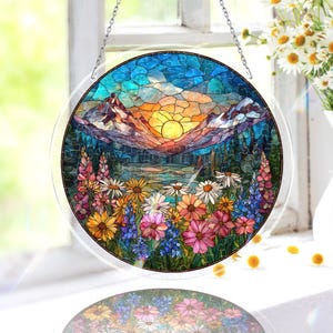 Peut inclure: Vitrail rond représentant un paysage de montagne avec un coucher de soleil, des arbres et des fleurs colorées. L'œuvre d'art est dans les tons de bleu, orange, jaune, rose et vert. La fenêtre est suspendue à une chaîne argentée.