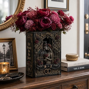 May include: A black rectangular vase shaped like a book, featuring a relief of two skeletons in wedding attire behind a gate. The text Till Death Do Us Part and S + J is visible. The vase is filled with a large arrangement of deep red and pink flowers.