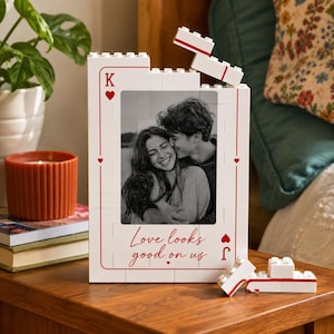 May include: A white rectangular picture frame constructed from interlocking building blocks, designed to resemble a playing card. It features a black and white photograph of a couple, red heart symbols, the letter K, and the text Love looks good on us.
