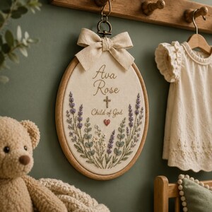 May include: An oval embroidery hoop wall hanging featuring the name Ava Rose, a small cross, and the text Child of God. The design includes embroidered lavender sprigs, small white flowers, and a red heart on cream fabric with a beige bow.