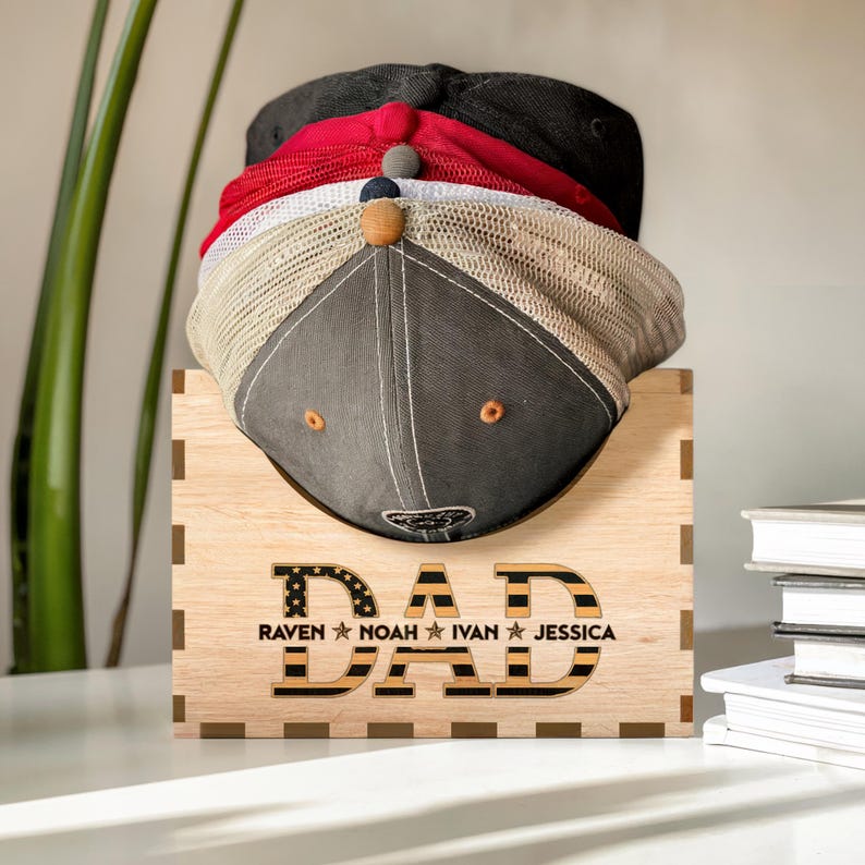 Puede incluir: Estante de sombreros de madera con la palabra "DAD" en letras negras y doradas con un patr&oacute;n de bandera estadounidense. El estante tiene los nombres "Raven", "Noah", "Ivan" y "Jessica" escritos debajo de la palabra "DAD".