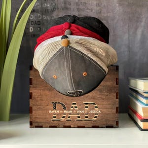 Puede incluir: Estante para sombreros de madera con la palabra "DAD" en letras grandes con un diseño de bandera estadounidense. El estante es marrón y tiene los nombres "Raven", "Noah", "Ivan" y "Jessica" escritos debajo de la palabra "DAD".