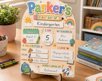 May include: A wooden arched sign for Parker's first day of kindergarten featuring colorful illustrations of a pencil, sun, rainbow, apple, globe, backpack, and rocket. The board includes text fields for date, teacher, age, favorites, and future career goals.