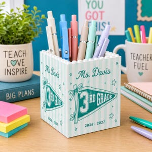 May include: A square white pen holder with a light teal striped pattern, featuring the text Ms. Davis, 3rd Grade, and 2026-2027. The design includes a teal pennant graphic with stars and bows. Several colorful pens are stored inside the container.