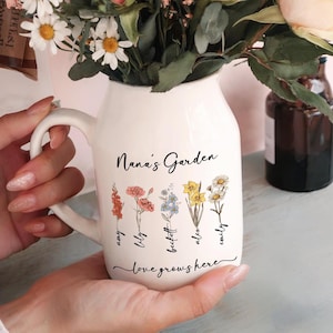May include: White ceramic pitcher with a handle, featuring the text "Nana's Garden" and a floral design with five flowers and names below each flower: "amy", "lily", "beckett", "alex", and "emily". The text "love grows here" is written below the flowers.