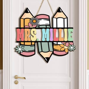 May include: A wooden door hanger featuring three colourful pencils and the text "MRS. MILLIE" in rainbow letters. The pencils have flowers on them.