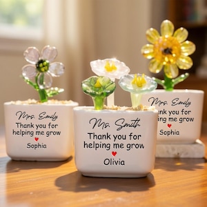 Pode incluir: Três vasos de cerâmica brancos com esculturas de flores de vidro. Cada vaso tem um design de flor diferente e inclui o texto "Obrigado por me ajudar a crescer" e um nome. As flores são em tons de amarelo, verde e transparente.
