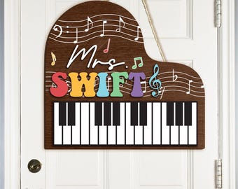 May include: A brown wooden piano-shaped door hanger with a white piano keyboard and the text "Mrs. SWIFT" in rainbow colors. Musical notes are printed on the brown background.