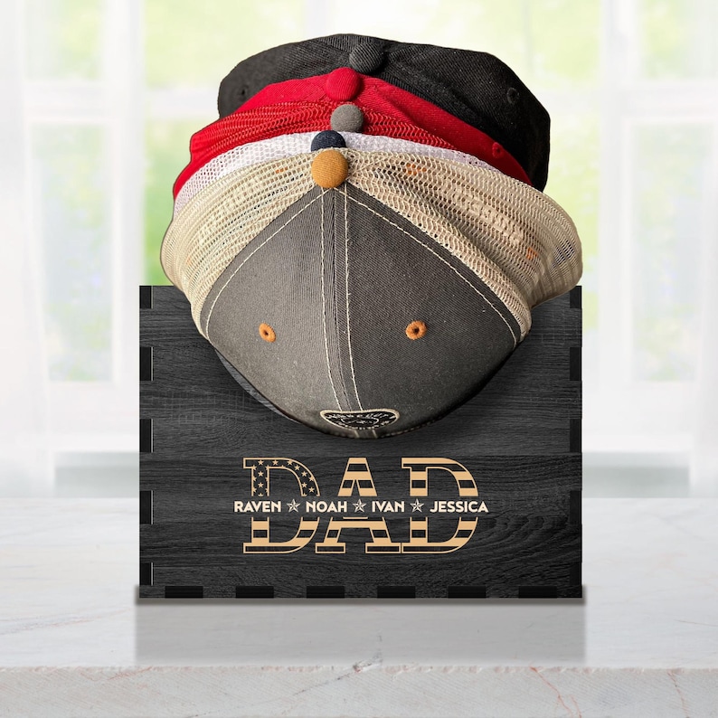 Puede incluir: Un perchero de sombreros de madera negro con la palabra "DAD" en letras doradas con un dise&ntilde;o de bandera estadounidense roja, blanca y azul. El perchero tiene cuatro ranuras para sombreros y los nombres "Raven, Noah, Ivan, Jessica" est&aacute;n impresos debajo de la palabra "DAD".