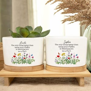 May include: Two white ceramic flower pots with wooden bases. The pots have a floral design and text that reads "Linda The only thing better than being your Mother is watching you be a Mom" and "Sophia The only thing better than being your Mother is watching you be a Mom".