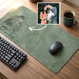 Op de afbeelding: Een groene muismat met een gegraveerd familieportret en de tekst "We love you Daddy". Een zwart computertoetsenbord en muis liggen op het houten bureau. Een kleine familiefoto staat in de rechterbovenhoek.