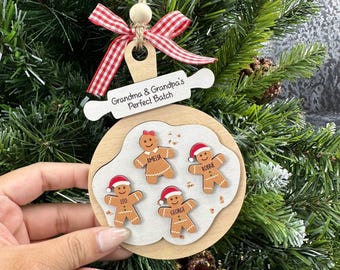 Puede incluir: Un adorno navideño de madera con un diseño de rodillo y lazo. El adorno presenta figuras de pan de jengibre con nombres y el texto "Grandma & Grandpa's Perfect Batch."