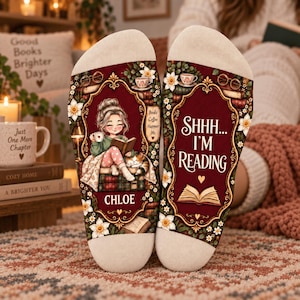 May include: A pair of cream-colored socks featuring a dark red central panel with ornate gold borders. The left sock shows an illustration of a person reading with a cat, while the right sock displays the text SHHH... I'M READING and an open book.