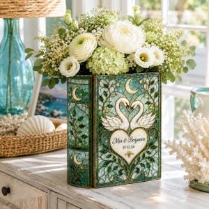 May include: A rectangular stained glass vase featuring a teal and green mosaic pattern with two white swans forming a heart shape. The center displays the text Mia & Benjamin and the date 07.12.26. The vase is filled with white flowers and greenery.