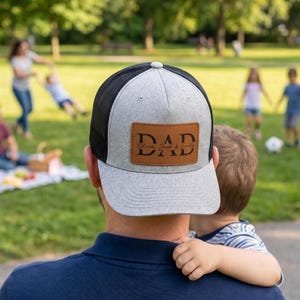 Puede incluir: Gorra de camionero gris y negra con un parche de cuero marrón que dice "DAD" en letras negras. El parche también incluye los nombres "Oliver, Jack & Emily". La gorra la lleva una persona que sostiene a un niño.
