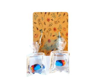 May include: A pair of novelty earrings featuring small, colorful fish encased in clear, plastic bags. The fish have blue, red, and yellow coloring. The earrings hang from silver-colored hooks and are displayed on a floral-patterned, brown card.