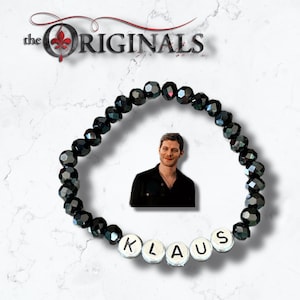 Puede incluir: Pulsera de cuentas negras con un dije plateado que dice "KLAUS" y una foto de un hombre con cabello oscuro y una camisa negra. La pulsera está sobre un fondo blanco con las palabras "the ORIGINALS" en rojo en la parte superior.