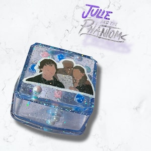 Può includere: Una scatola di resina blu trasparente con glitter e un adesivo della band "Julie and the Phantoms".