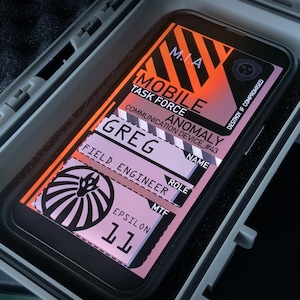 May include: A smartphone with a custom interface, displayed inside a gray protective case. The screen shows text including "MOBILE TASK FORCE" and "GREG", with a graphic design. The case is open, revealing the phone's screen.