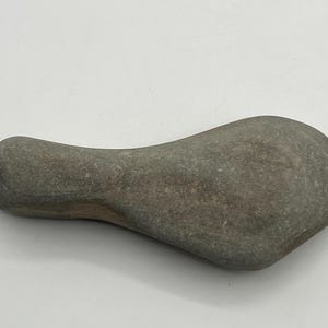 May include: A smooth, gray, elongated river stone with a rounded, bulbous end and a wider, flattened section. The surface of the rock features natural variations in color and texture, displaying a matte finish with subtle speckling throughout.