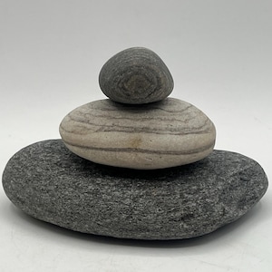 May include: A stack of three smooth, gray stones of varying sizes. The bottom stone is the largest, followed by a medium-sized stone with horizontal stripes, and a small, round stone on top. The stones are arranged on a white background.