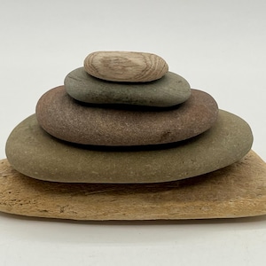 May include: A stack of five smooth, flat stones arranged in a pyramid shape. The base is a large, light tan, textured stone, topped by a medium-sized olive-green stone, a larger reddish-brown stone, a smaller gray stone, and a light tan top stone.