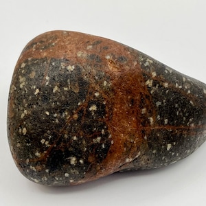 May include: A smooth, rounded stone with a speckled, dark gray base and a prominent band of reddish-brown across the top. The stone's surface is dotted with small, white flecks, creating a textured appearance. The stone is a natural, earthy item.