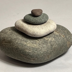 May include: A stack of five smooth, rounded stones in varying sizes and colors. The largest stone is a mottled gray, supporting a white stone, a green stone, and a small brown stone at the top. The stones are arranged in a balanced, natural composition.
