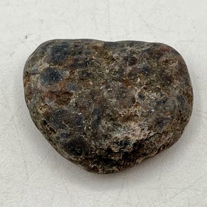 May include: A heart-shaped, natural stone with a rough, textured surface. The stone features a mix of dark brown, black, and lighter tan colors, creating a mottled appearance. The overall shape is rounded and smooth, suggesting it has been worn by water.