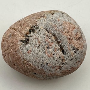 May include: A smooth, rounded river stone featuring a mottled texture with a blend of pink, gray, and tan mineral flecks. The surface displays a natural, weathered appearance with a small, dark, irregular crevice running across the center.