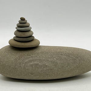 May include: A balanced stone sculpture featuring a large, smooth, oval-shaped base rock in a light brown hue. Upon it, a stack of seven smaller, rounded stones of varying sizes and shades of gray and brown are carefully arranged, creating a zen-like aesthetic.
