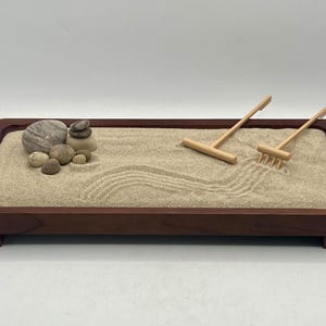 May include: A miniature Zen garden in a dark wood tray. The sand is raked with wave patterns. A small pile of stones and two wooden rakes are included. The tray is rectangular with a raised edge.