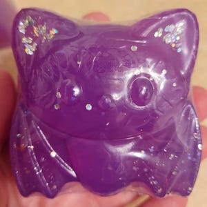 May include: A purple bat-shaped resin figure with glitter accents. The bat has large, round eyes, pointed ears, and outstretched wings. The figure is held in a hand, and the background is blurred.