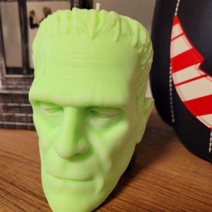 May include: A lime green candle shaped like Frankenstein's monster's head. The candle has detailed facial features, including a prominent brow, nose, and defined lips. The candle is a unique decorative item for Halloween or horror enthusiasts.