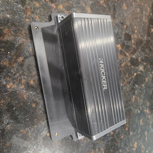 May include: A black Kicker KEY200.4 amplifier mounted on a black bracket. The amplifier has a ribbed design and the Kicker logo is visible. The bracket has screw holes for mounting.