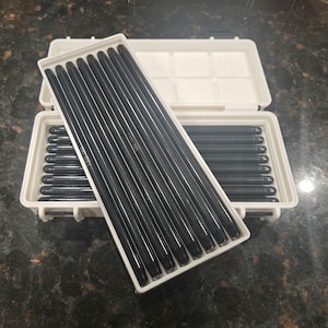 May include: A set of black rods in a white plastic storage case. The rods are arranged in rows within the case. The case is open, revealing the contents. The background is a dark, speckled surface.
