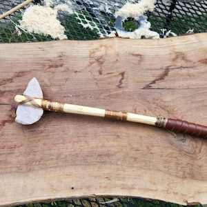 May include: A handmade stone axe with a light-colored wooden handle. The axe head is a light gray stone, secured with wrapped twine. The handle transitions to a brown leather grip.