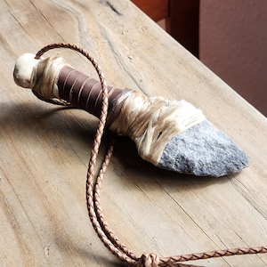 Puede incluir: Un cuchillo de piedra hecho a mano con un cordón de cuero trenzado. El mango está envuelto en cuero marrón y fibras naturales. La hoja es de piedra gris y el cordón es de color marrón claro.