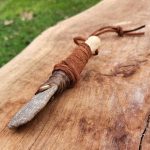 Puede incluir: Un cuchillo de piedra hecho a mano con un mango envuelto en cuero marrón y un detalle de hueso. La hoja es de color gris claro y el mango está envuelto con cuero marrón más oscuro. Una cuerda de cuero marrón está unida al mango.