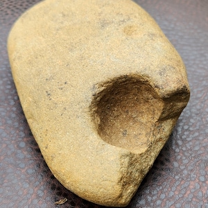 Op de afbeelding: Een bruin-gele steen met een afgeronde vorm en een cirkelvormige uitsparing. De steen heeft een gladde, versleten textuur, wat suggereert dat het een natuurlijk artefact is. Het kan een gereedschap of een decoratief item zijn.