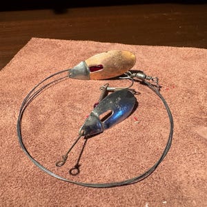 May include: Two fishing lures with metal and wood components, connected by a wire. One lure has a wooden body with a red accent, the other is metallic blue. The lures are on a brown, textured surface.