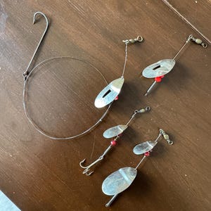 May include: Fishing lures and a wire leader are arranged on a wooden surface. The lures have silver-colored, oval-shaped blades with red beads and hooks. The wire leader has a large hook at one end.