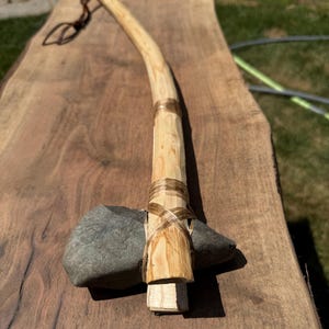 Puede incluir: Un hacha de piedra artesanal con una cabeza de piedra gris oscuro de forma irregular, sujeta firmemente a un mango de madera clara y curvada con un material de unión fino y translúcido. Un cordón de cuero marrón fino está en el extremo.