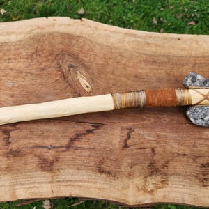 Puede incluir: Un hacha de piedra artesanal con un mango de madera clara. El mango está envuelto con cuerda y cuero marrón. La cabeza del hacha es de piedra gris. El hacha se exhibe sobre una superficie de madera.