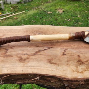 Puede incluir: Un hacha de piedra artesanal con un mango de madera envuelto en cuero marrón. La cabeza del hacha es de piedra gris y el mango es de madera clara. El hacha se exhibe sobre una superficie de madera.