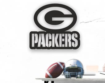 Puede incluir: Un letrero de metal negro con el logotipo de los Green Bay Packers, una gran "G" verde sobre la palabra "PACKERS" en blanco. Un balón de fútbol marrón y un casco plateado están sobre una superficie blanca.