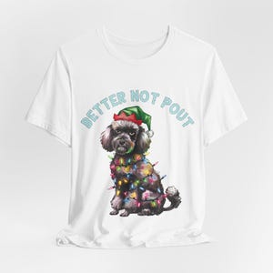 May include: White t-shirt featuring a black and gray dog wearing a green elf hat and wrapped in colorful Christmas lights. The text "BETTER NOT POUT" is printed above the dog in light blue letters.