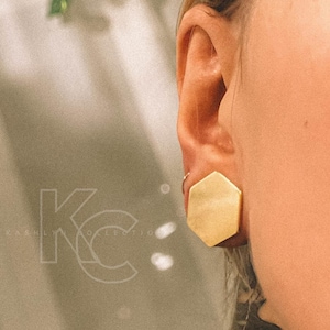 May include: A gold hexagonal earring with a smooth, polished finish. The earring is worn in the upper lobe of a person's ear.