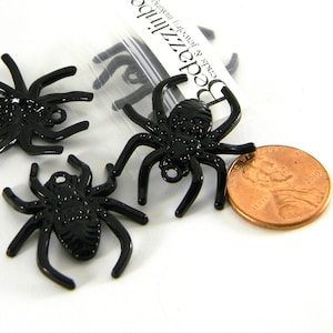 Op de afbeelding: Zwarte plastic spinnencharms, ongeveer de grootte van een Nederlandse cent. De spinnen hebben een getextureerd lichaam en acht poten. De charms hebben een kleine lus aan de bovenkant om op te hangen.